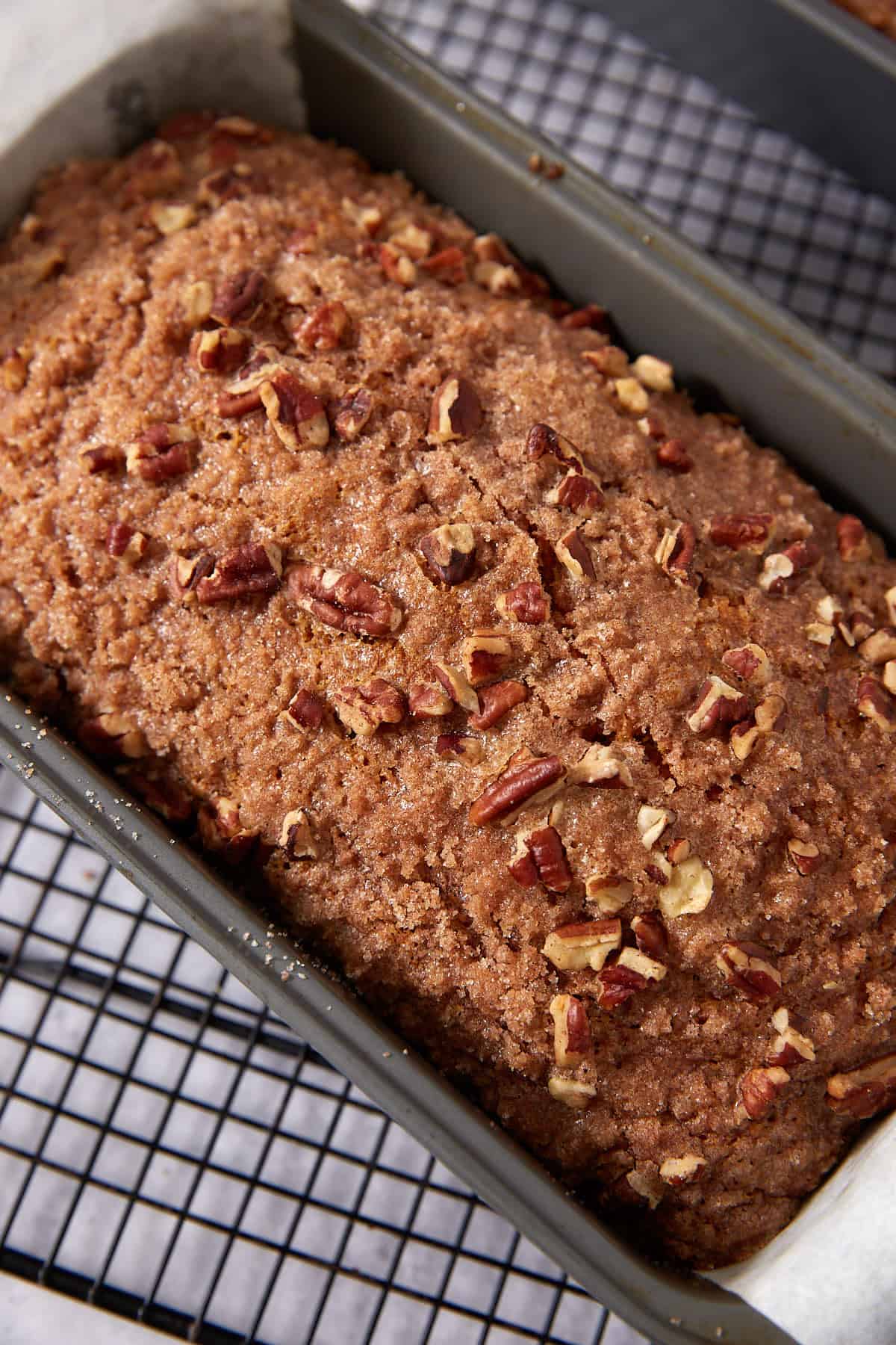 A freshly baked loaf of bread topped with chopped pecans sits in a metal pan on a cooling rack.