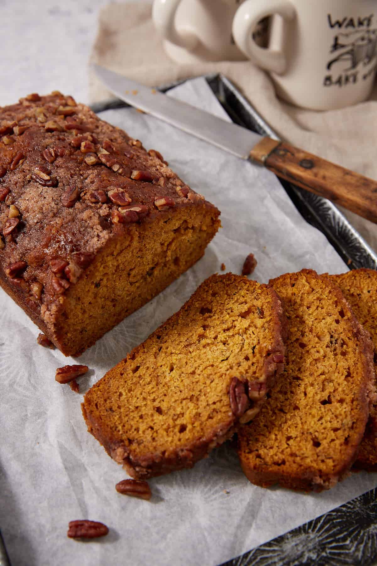A loaf of pumpkin bread topped with pecans sits on parchment paper, with two slices cut. A knife and two mugs are in the background.