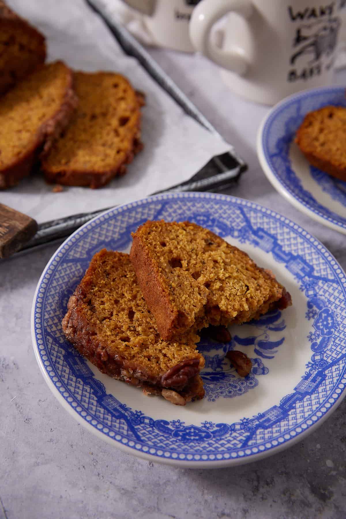 Two slices of pumpkin bread on a blue and white patterned plate, with more slices and coffee mugs in the background on a gray surface.