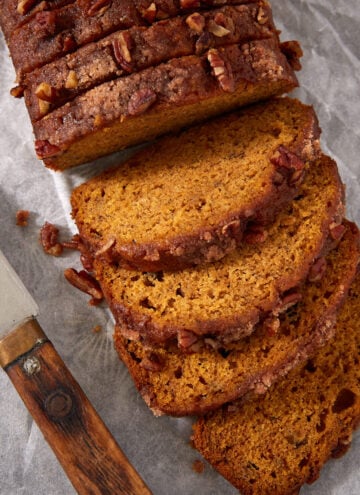 A loaf of sliced pumpkin bread with pecans on top sits on parchment paper next to a knife with a wooden handle.