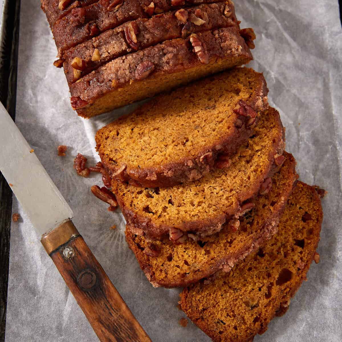 A loaf of sliced pumpkin bread with pecans on top sits on parchment paper next to a knife with a wooden handle.