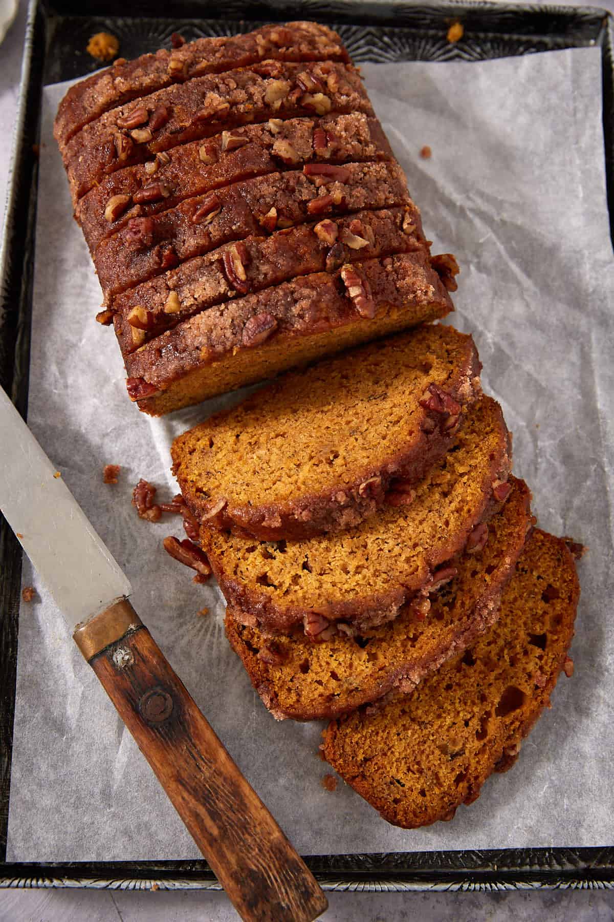 A loaf of pumpkin bread with a crumbly pecan topping is sliced on a baking tray lined with parchment paper. A wooden-handled knife rests beside the bread.