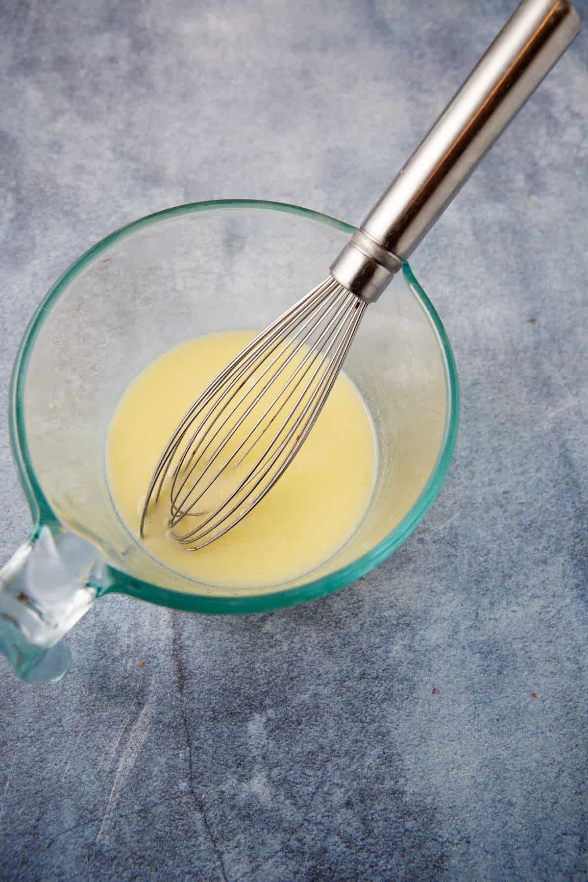 A glass measuring cup with a metal whisk inside, containing a small amount of pale yellow liquid, sits on a textured gray-blue surface.