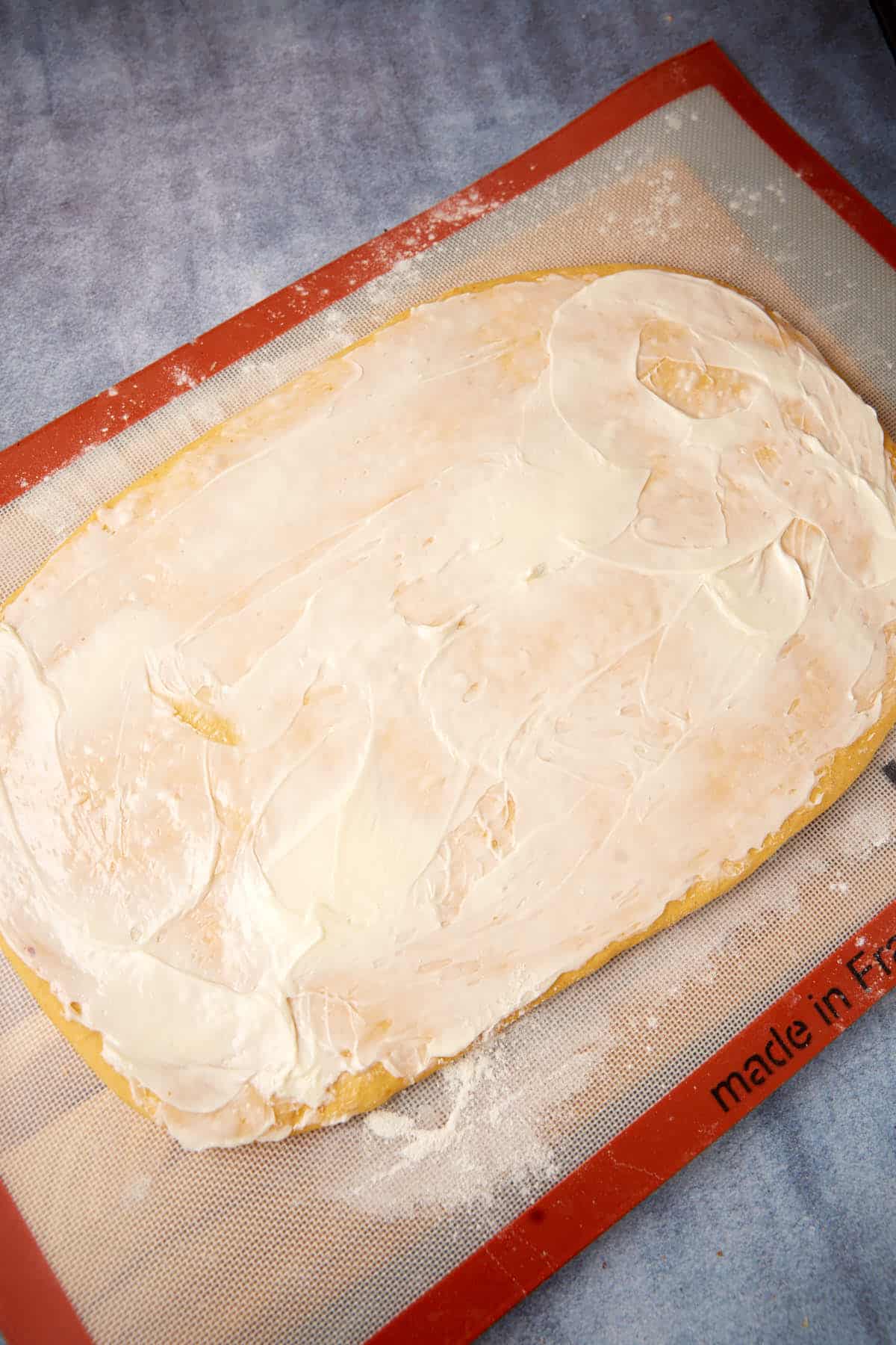 A rolled-out sheet of dough topped with a layer of cream cheese sits on a baking mat on a blue countertop.