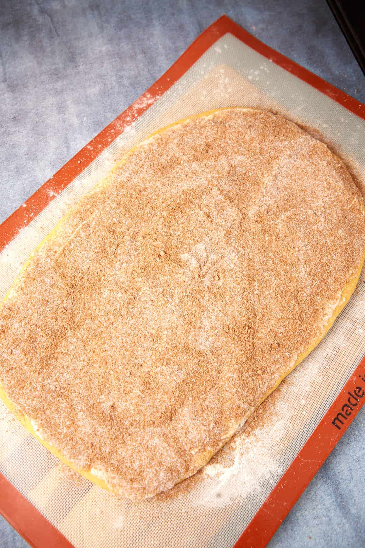 An oval-shaped dough is covered with a cinnamon-sugar mixture, resting on a silicone baking mat on a blue countertop.