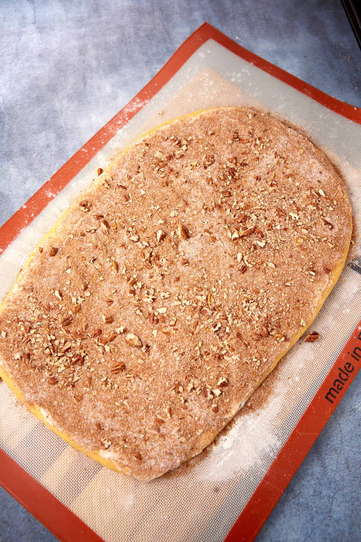 Raw dough rolled out on a silicone baking mat, topped with a layer of cinnamon, sugar, and chopped pecans, ready for baking.