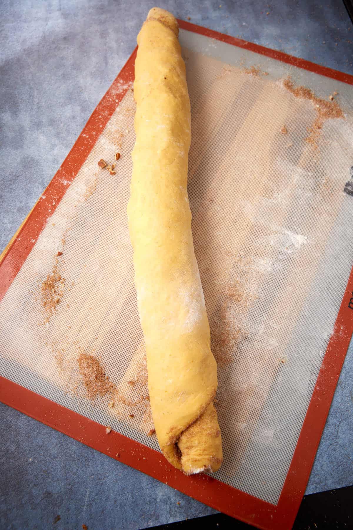 A rolled dough log filled with cinnamon on a silicone baking mat, ready to be sliced for making cinnamon rolls. Flour and cinnamon sugar are scattered on the mat.