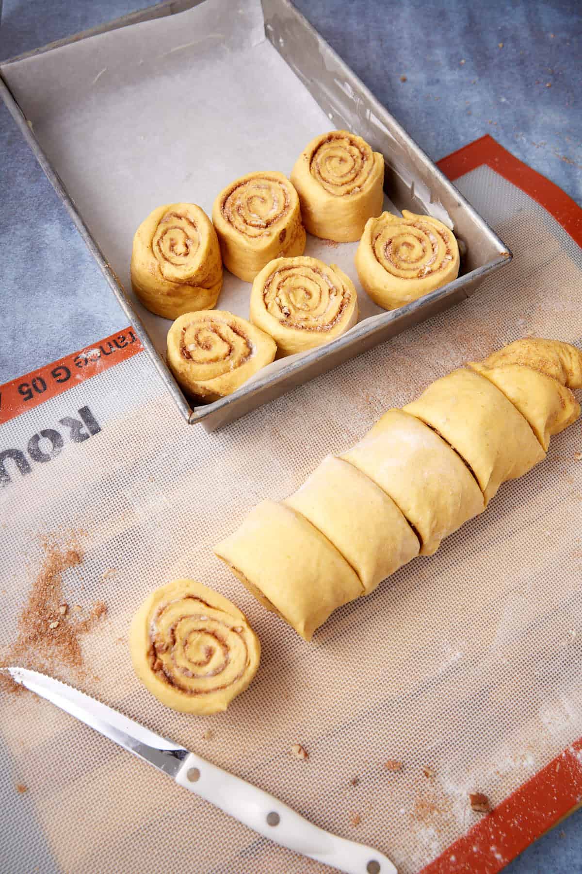 Unbaked cinnamon rolls being cut and placed in a metal baking pan; a knife and baking mat are nearby, and dough swirls are visible.