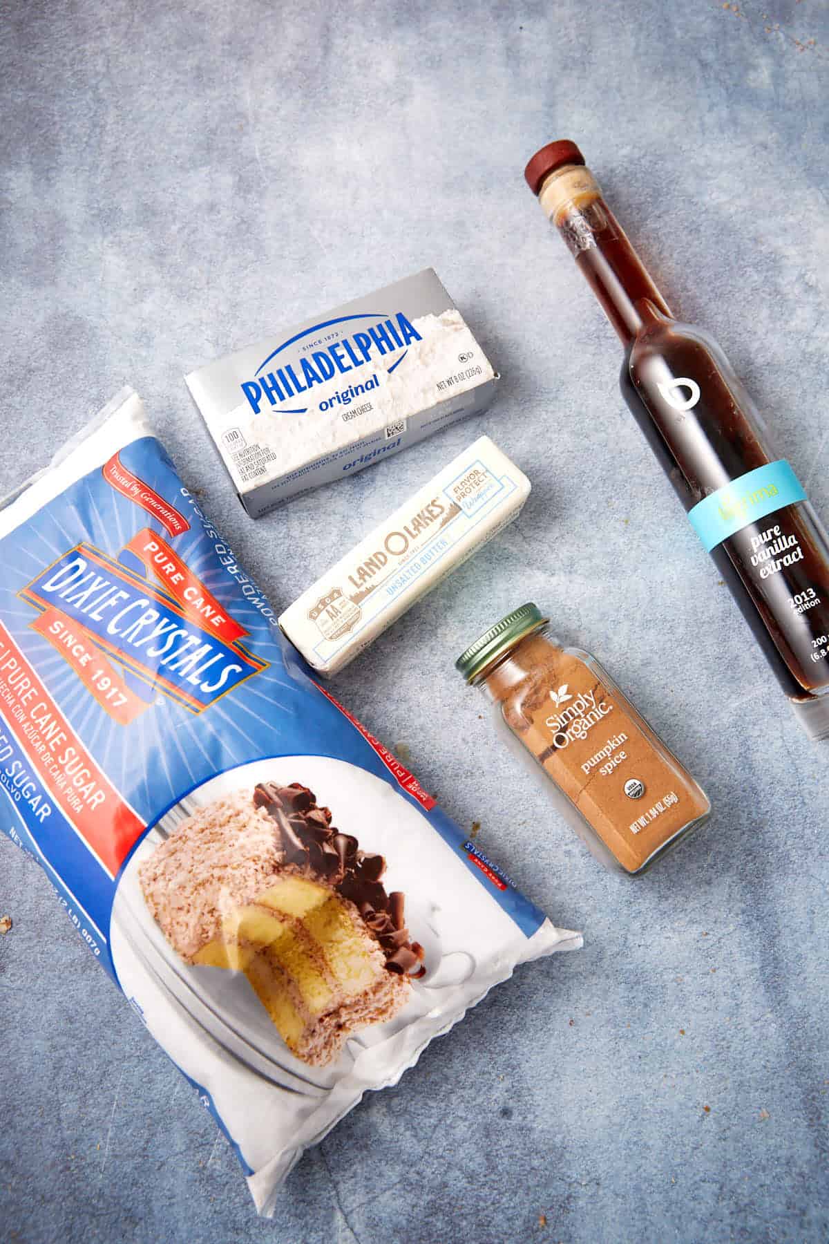 A bag of sugar, cream cheese, butter, ground cinnamon, and a bottle of vanilla extract are arranged on a blue textured surface.