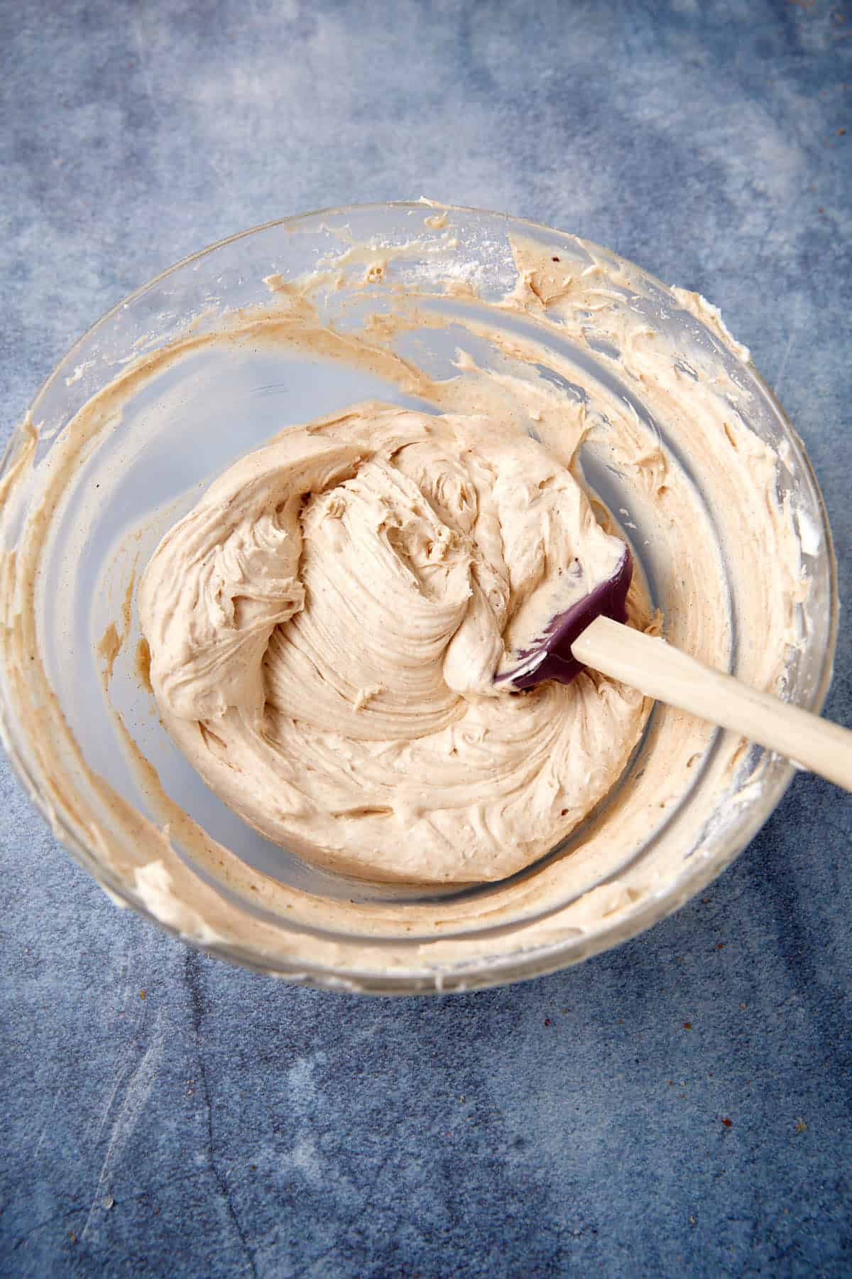 A glass bowl filled with creamy, light brown cake batter sits on a gray surface. A purple spatula with a wooden handle rests inside the bowl, coated with the batter.