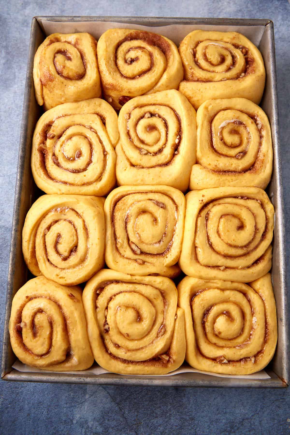 A metal baking pan filled with twelve unbaked cinnamon rolls arranged in neat rows on a grey surface.