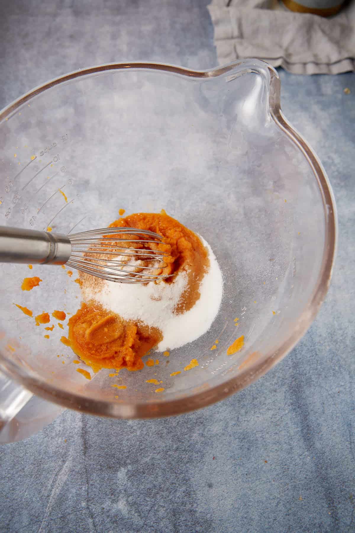 A clear mixing bowl with pumpkin puree, sugar, and spices being whisked together on a blue countertop.