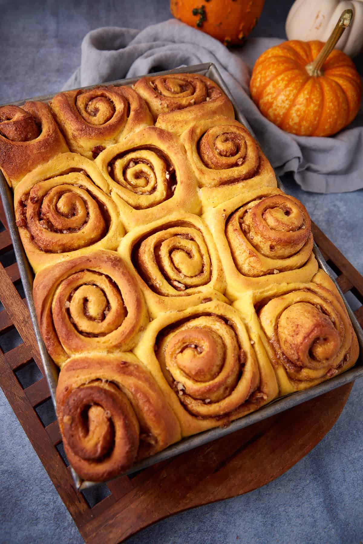 A tray of freshly baked pumpkin cinnamon rolls sits on a wooden trivet, with two small pumpkins and a gray cloth in the background.