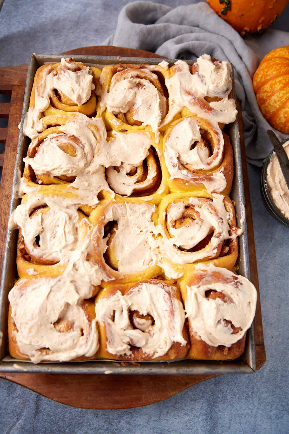 A baking tray filled with twelve frosted cinnamon rolls sits on a blue surface, with a bowl of icing, a gray cloth, and two small pumpkins nearby.