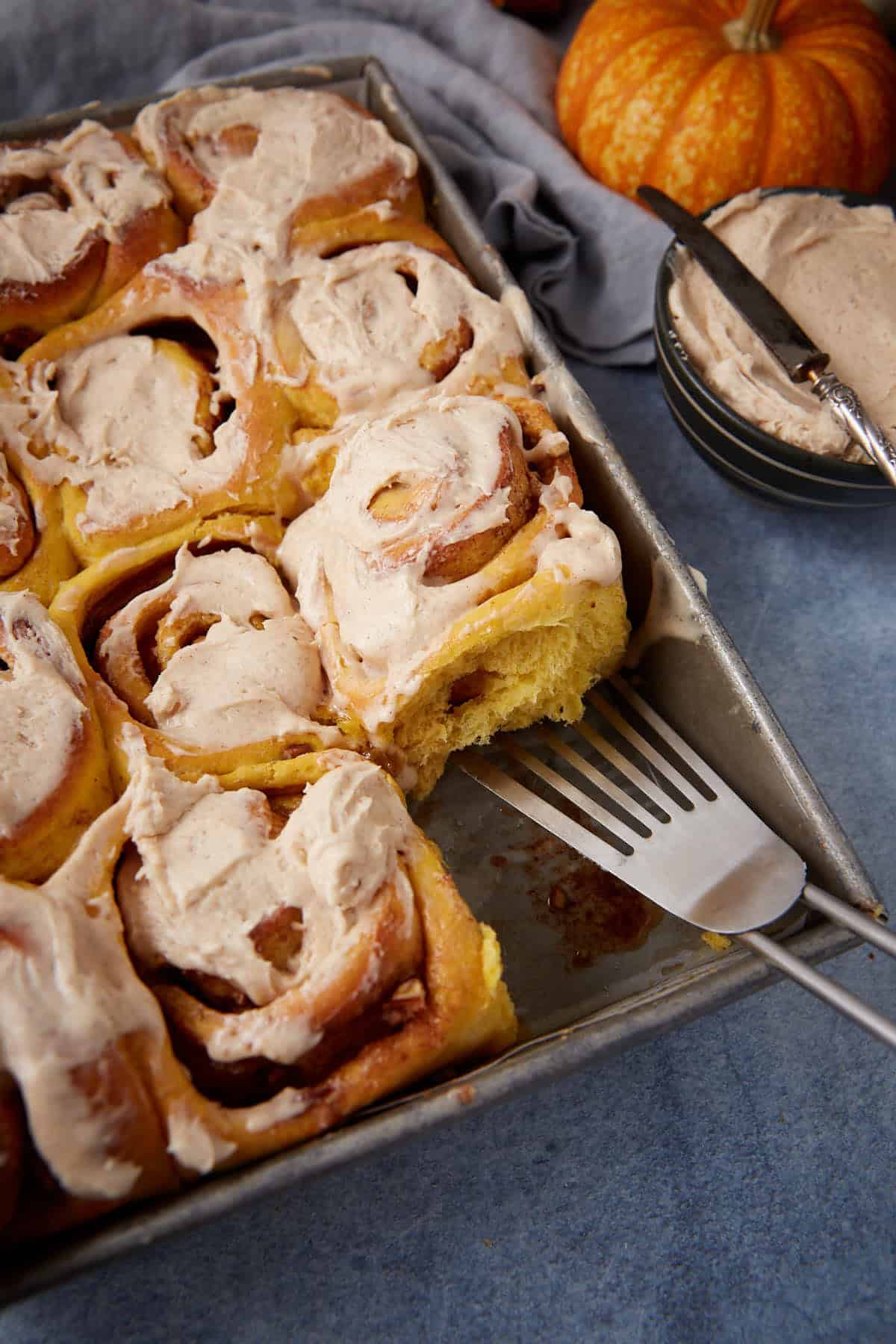 A tray of frosted cinnamon rolls, one being lifted out with a spatula. A bowl of extra frosting with a knife, a pumpkin, and a gray cloth are nearby on a blue surface.