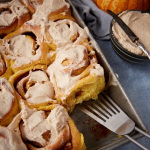 A tray of frosted cinnamon rolls, with one roll being lifted out by a spatula. A bowl of extra frosting and a butter knife sit nearby.