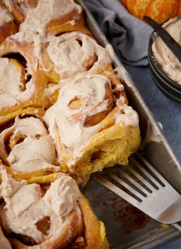 A tray of frosted cinnamon rolls, with one roll being lifted out by a spatula. A bowl of extra frosting and a butter knife sit nearby.