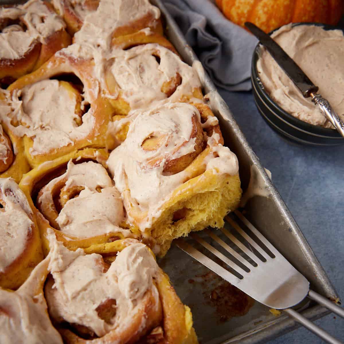 A tray of frosted cinnamon rolls, with one roll being lifted out by a spatula. A bowl of extra frosting and a butter knife sit nearby.