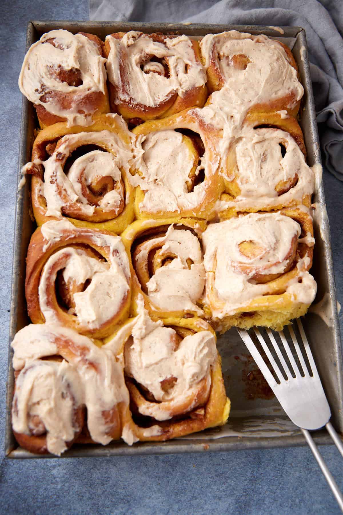 A metal pan filled with nine frosted cinnamon rolls, one of which is slightly lifted by a metal spatula, sits on a blue surface with a gray cloth nearby.