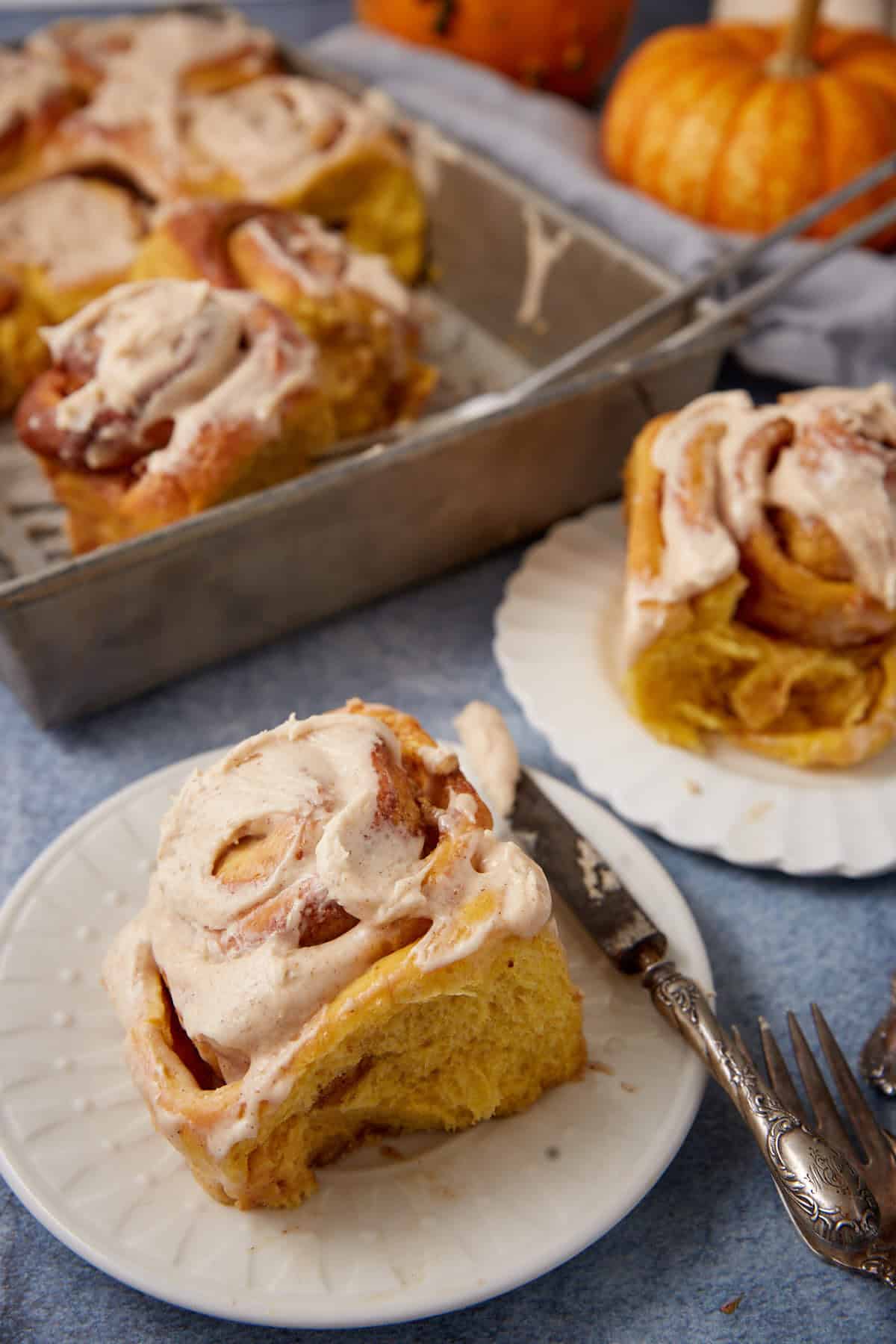 A frosted pumpkin cinnamon roll sits on a white plate with a fork, while more rolls are in a metal baking dish and another plate. Small pumpkins are in the background.