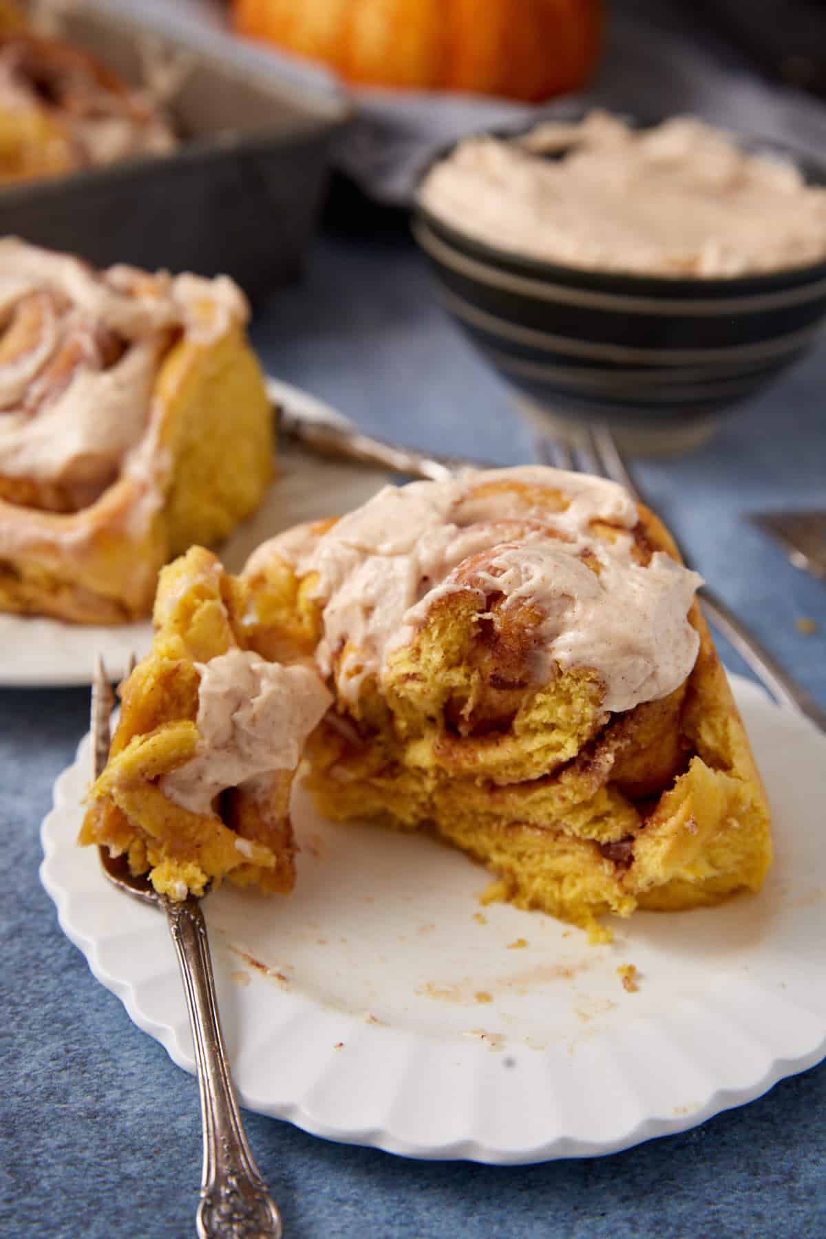 A pumpkin cinnamon roll with creamy frosting sits on a white plate, partially eaten with a fork. Another roll and a bowl of frosting are in the background on a blue surface.