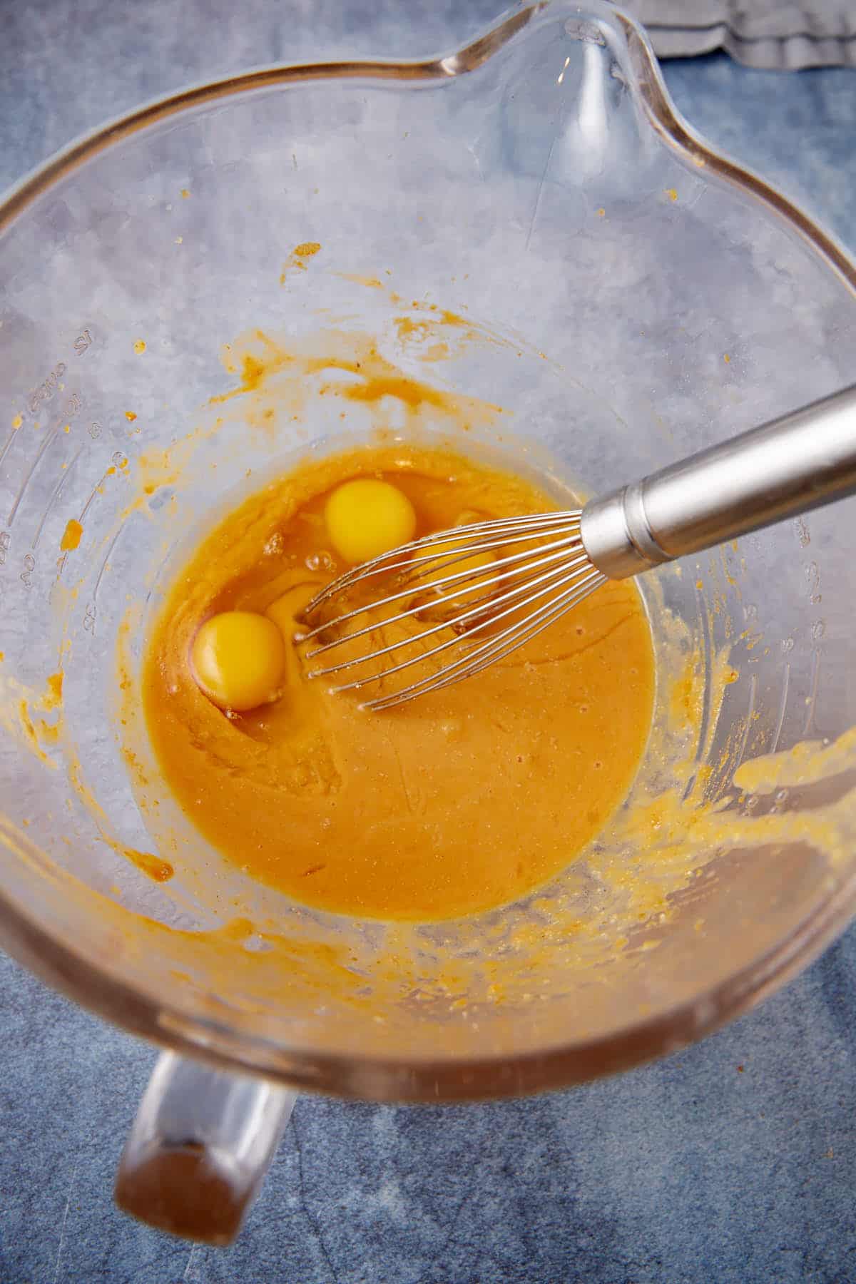 A whisk rests in a glass mixing bowl containing two raw eggs and a yellow batter on a blue countertop.