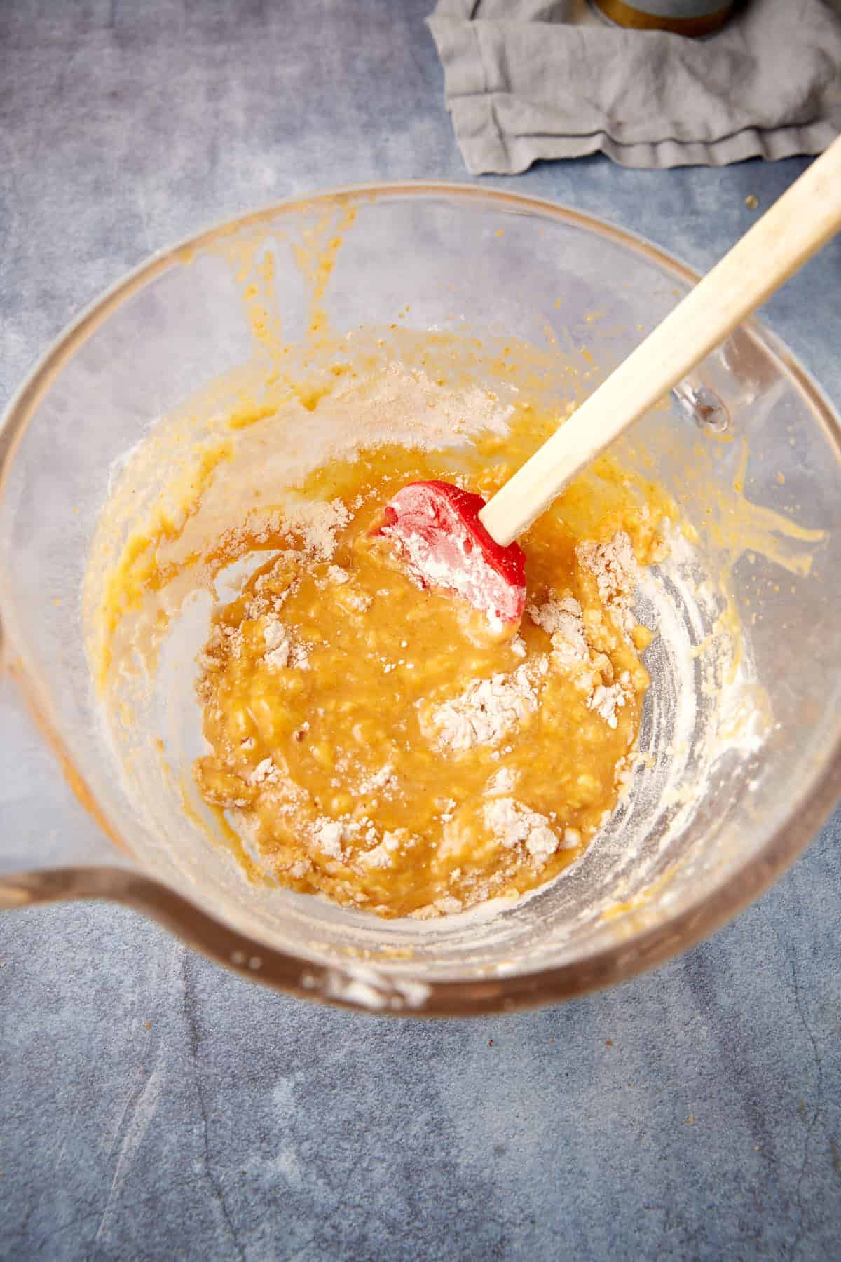 A glass mixing bowl with a red spatula stirring a thick, partially mixed batter. Some flour is still visible on top. The bowl sits on a blue-gray countertop with a gray cloth in the background.