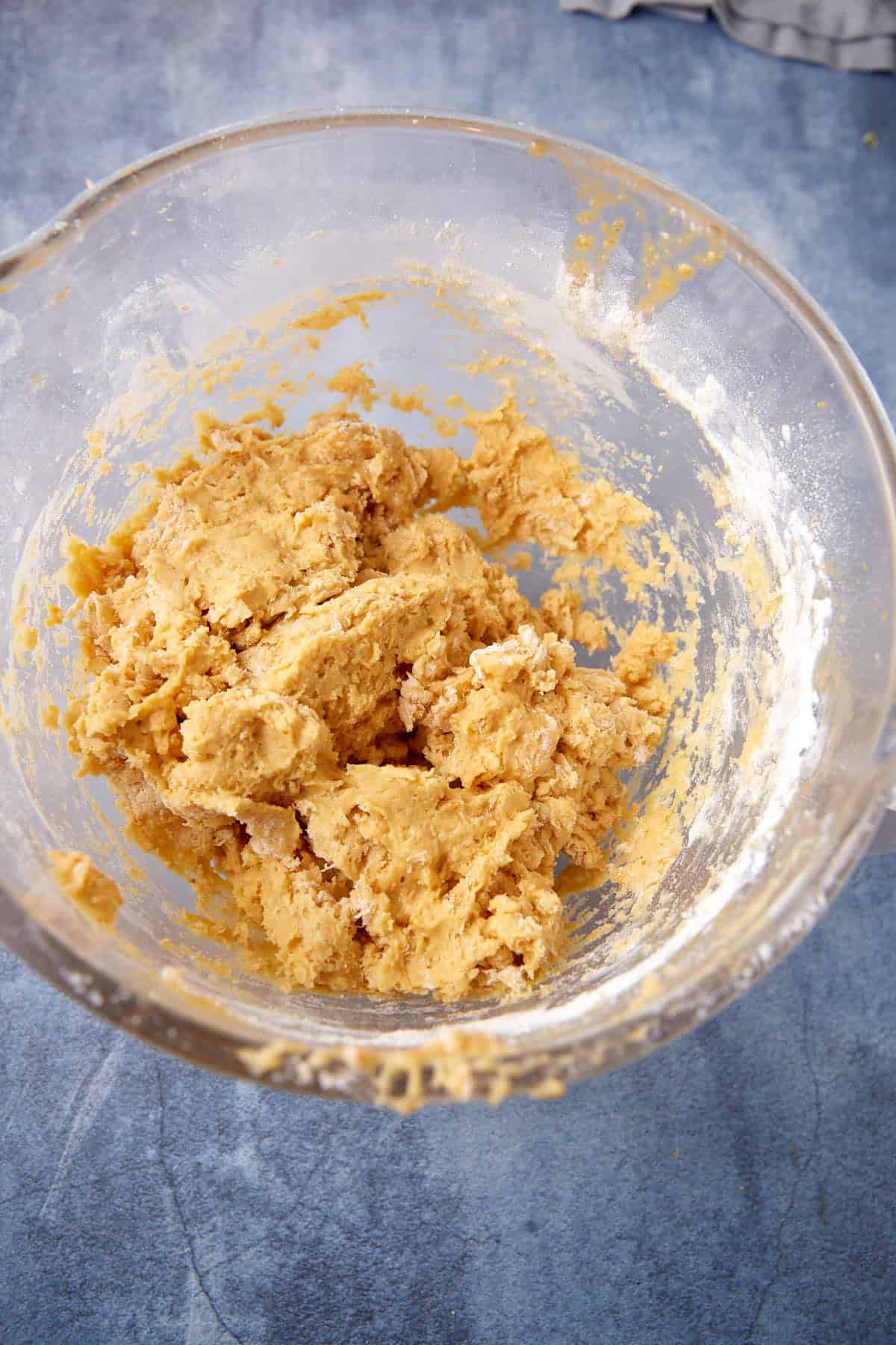 A clear glass bowl filled with thick, mixed cookie dough sits on a blue countertop. Some flour is stuck to the sides of the bowl.
