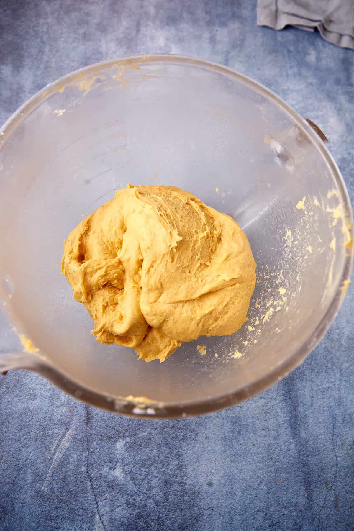 A ball of cookie dough sits in a clear mixing bowl on a blue countertop, with a gray cloth partly visible in the corner.