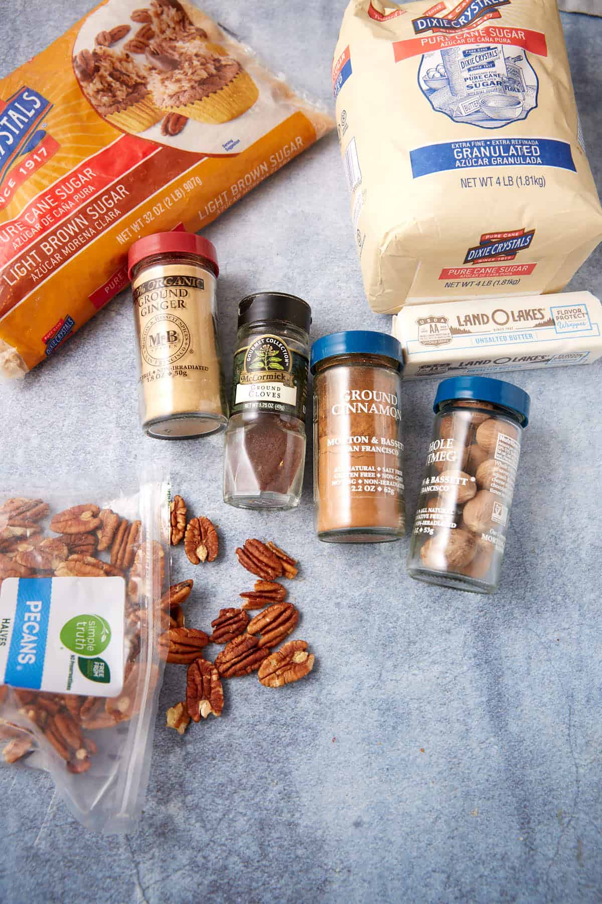 Bags of brown and granulated sugar, pecans, a stick of butter, and jars of ground ginger, cinnamon, cloves, and whole nutmeg laid out on a blue surface.