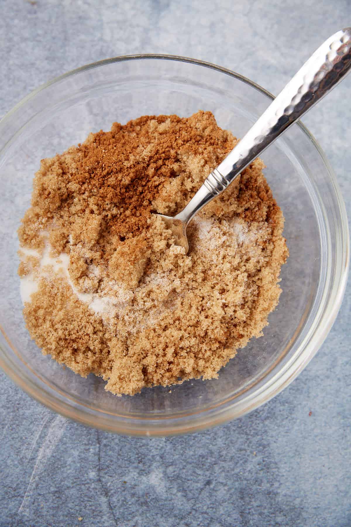A glass bowl filled with brown sugar, cinnamon, and white sugar, with a metal spoon resting inside, on a light blue textured surface.