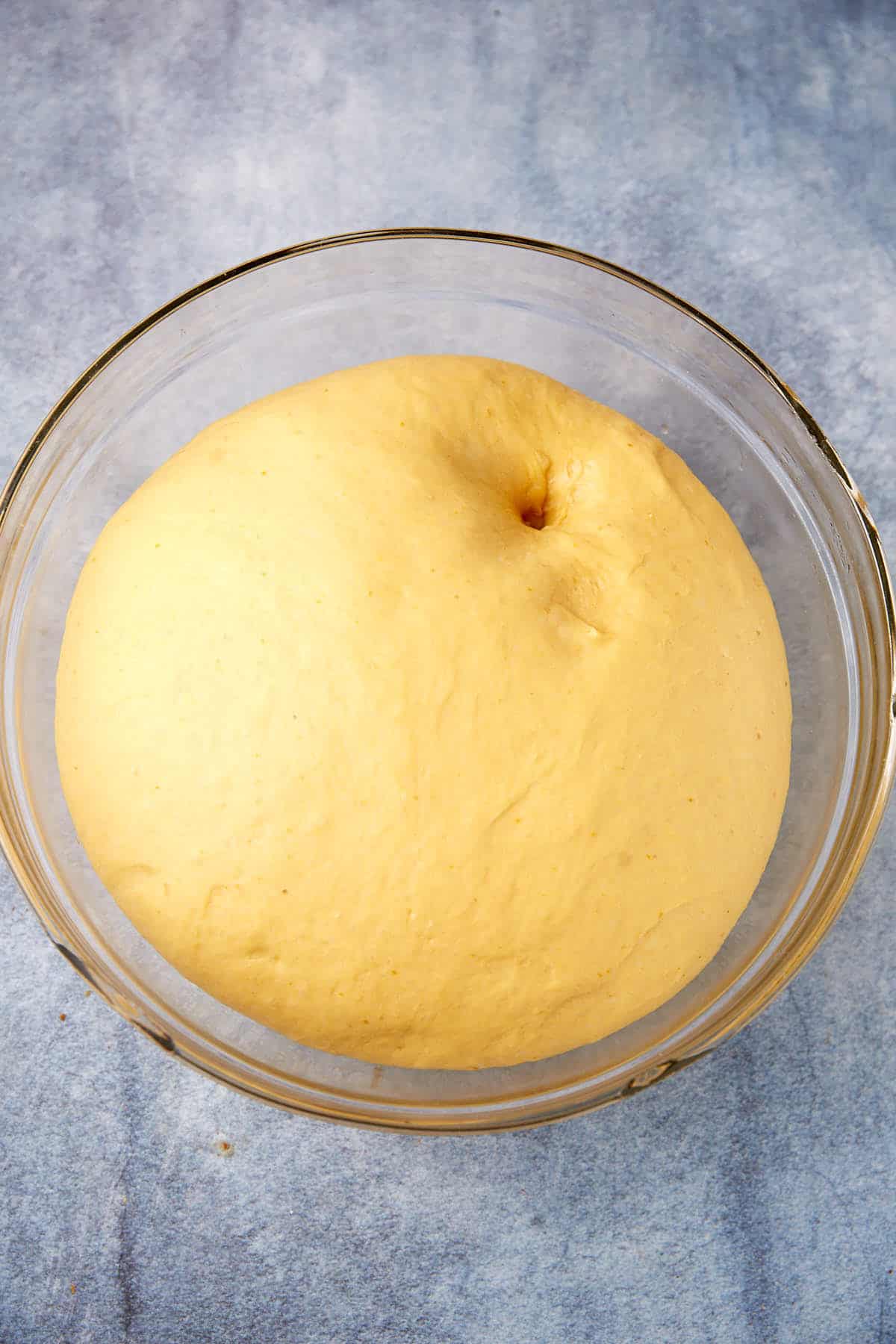 A large, risen ball of dough with a finger indentation sits in a clear glass bowl on a blue-gray countertop.