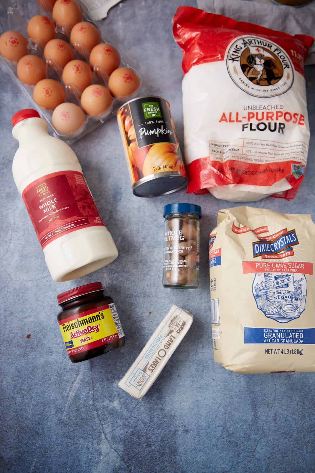 Baking ingredients on a blue surface: a carton of eggs, whole milk, canned pumpkin, ground cinnamon, all-purpose flour, granulated sugar, dry yeast jar, and a stick of unsalted butter.