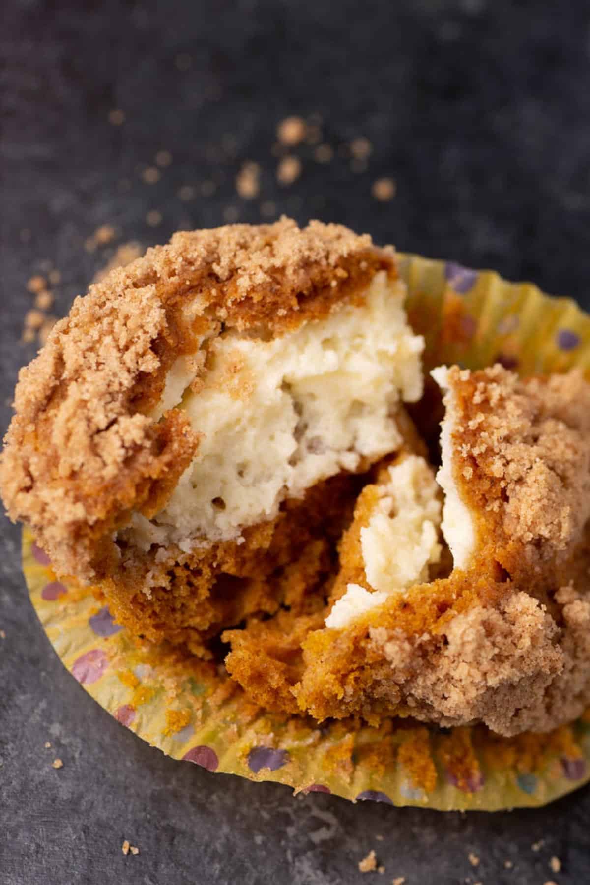 A close-up of a muffin with a crumb topping, split open to reveal a creamy filling inside, sitting on a colorful muffin liner with crumbs scattered around.