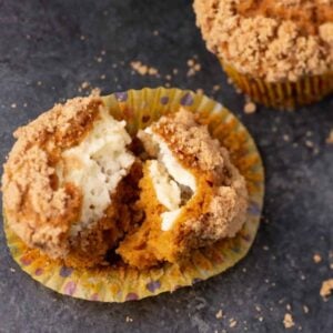 Pumpkin muffin with crumb topping split open, revealing a creamy filling inside. Another whole muffin is partially visible in the background on a dark surface with crumbs scattered around.