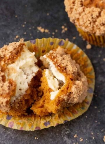 Pumpkin muffin with crumb topping split open, revealing a creamy filling inside. Another whole muffin is partially visible in the background on a dark surface with crumbs scattered around.