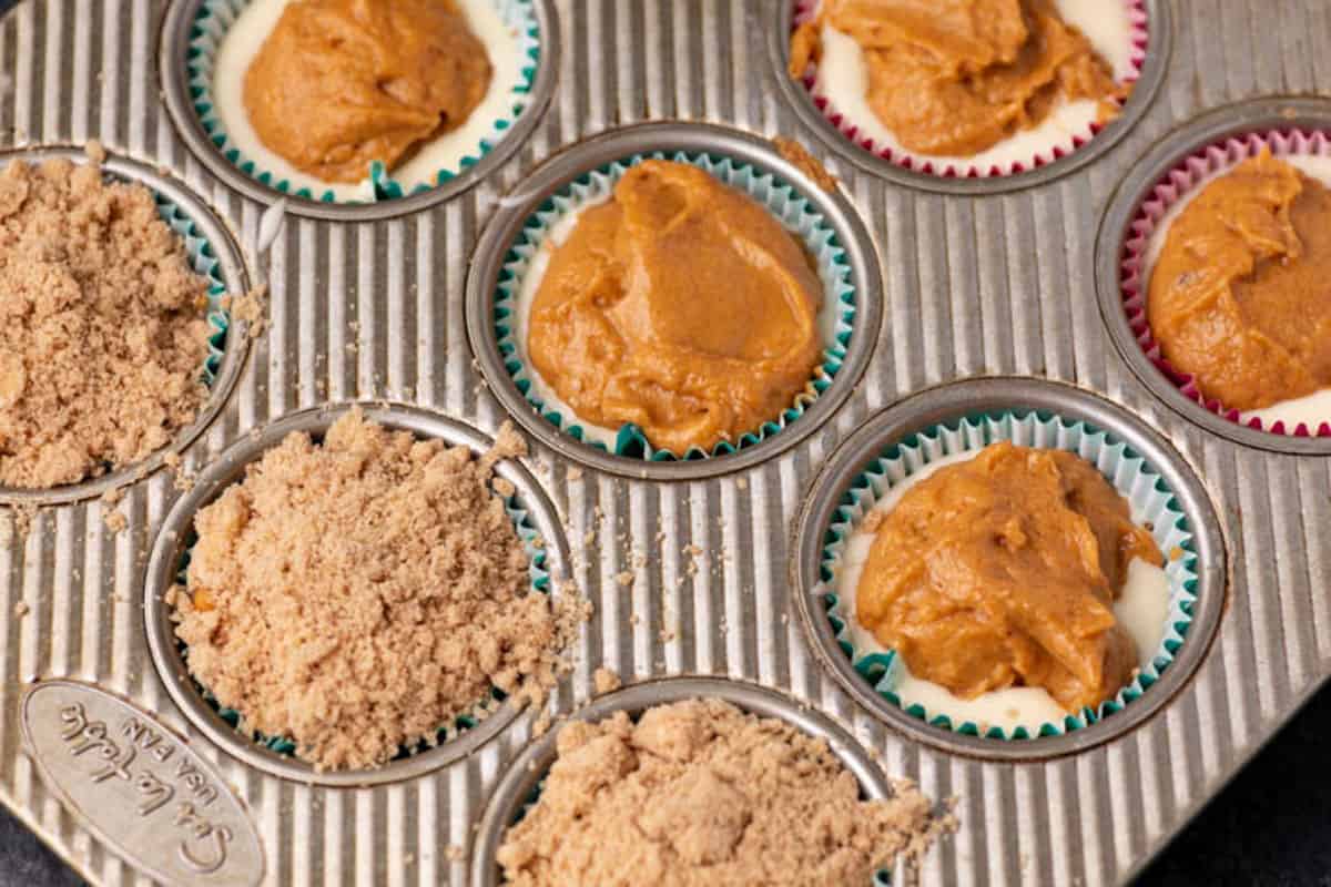 A muffin tin holds cupcake liners filled with batter. Some have a brown cinnamon mixture on top, while others are covered with brown sugar crumb topping, ready to be baked.