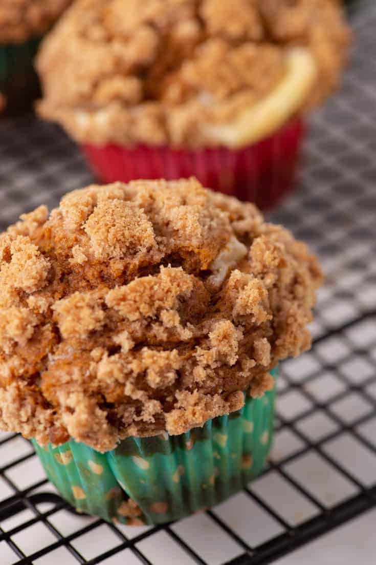 A close-up of a cinnamon streusel muffin in a green polka-dot wrapper, cooling on a black wire rack, with another muffin in the background.
