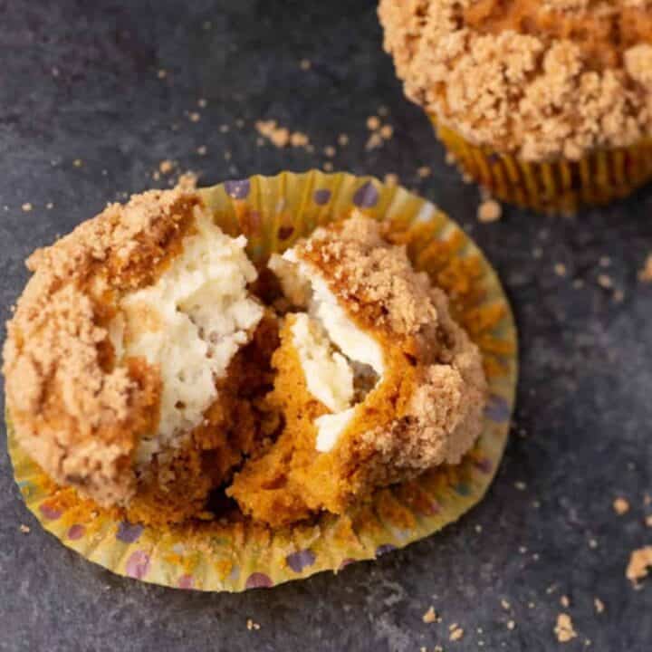 Pumpkin muffin with crumb topping split open, revealing a creamy filling inside. Another whole muffin is partially visible in the background on a dark surface with crumbs scattered around.