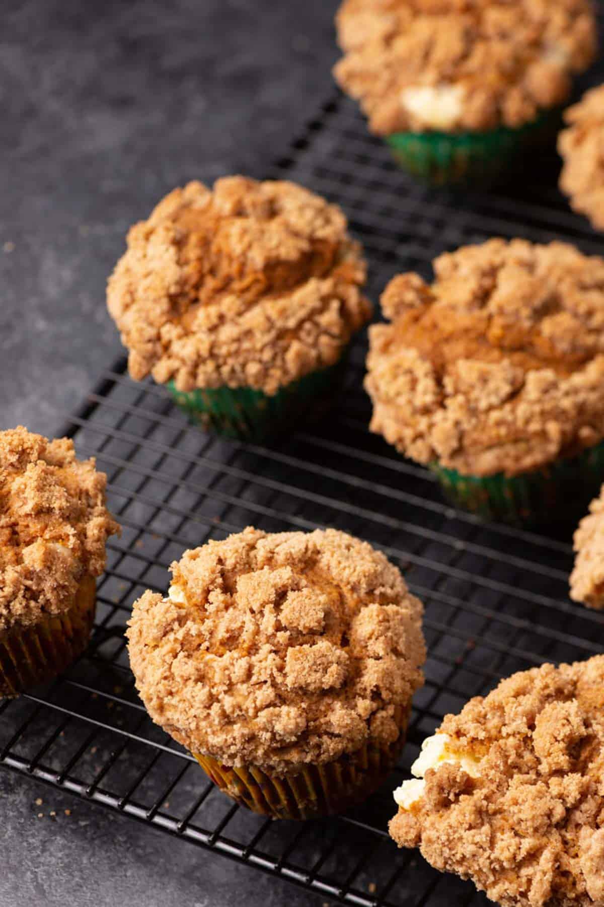 Seven crumb-topped muffins cool on a black wire rack, with a dark tabletop background. The muffins have a golden-brown, crumbly streusel topping.