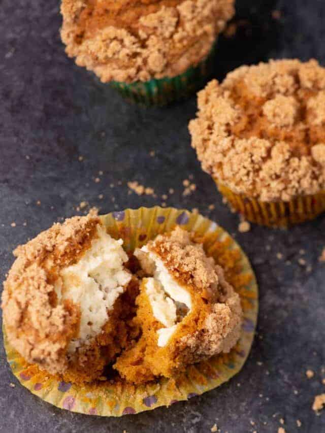 Three crumb-topped muffins sit on a dark surface. One muffin is split open, revealing a creamy filling inside. Crumbs and a colorful paper liner are visible around the muffins.