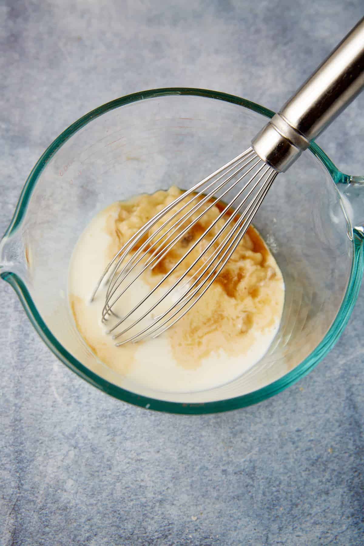 A metal whisk rests in a glass bowl containing milk, mashed bananas, and vanilla extract, ready to be mixed on a light blue textured surface.