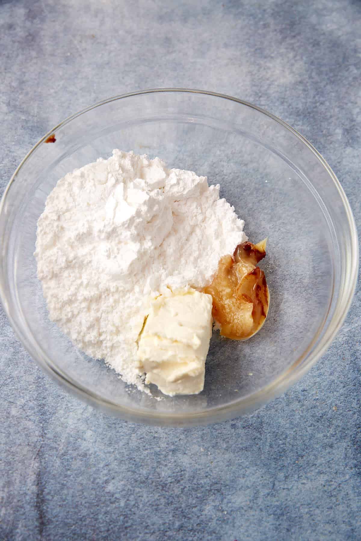 A glass bowl containing powdered sugar, a chunk of cream cheese, and a small portion of roasted garlic on a blue surface.