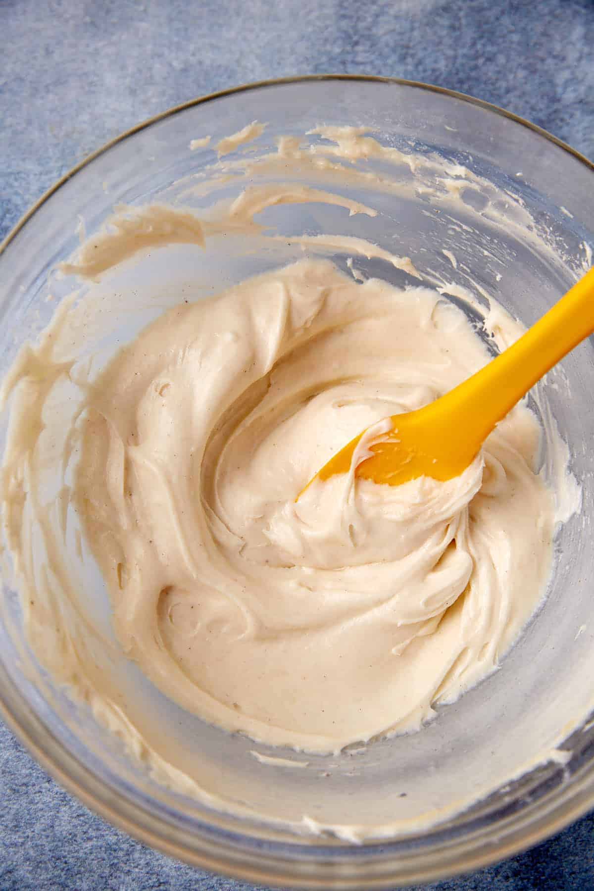 A glass bowl filled with creamy beige batter being mixed with a yellow spatula, set on a blue surface.
