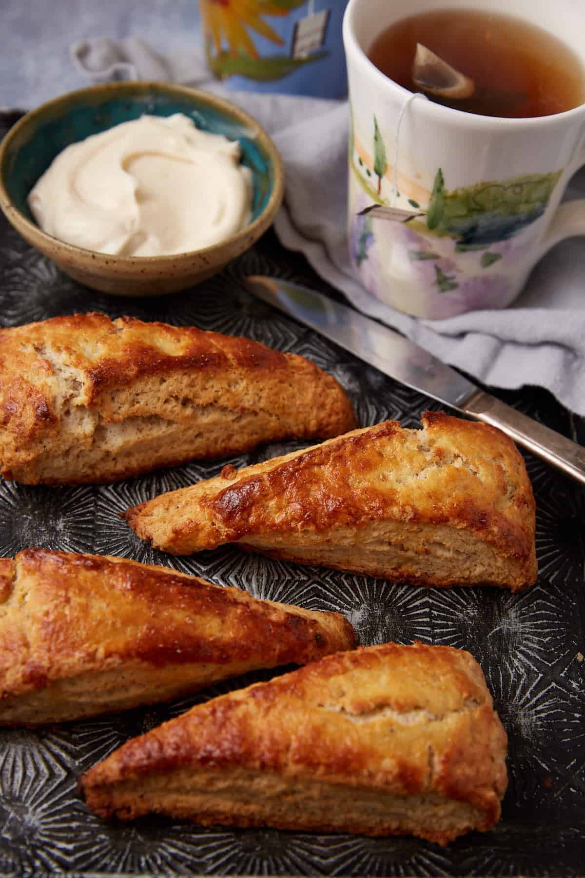 Four golden-brown scones on a patterned tray with a bowl of cream, a knife, and a floral mug of tea with a tea bag. A gray cloth is partially visible in the background.