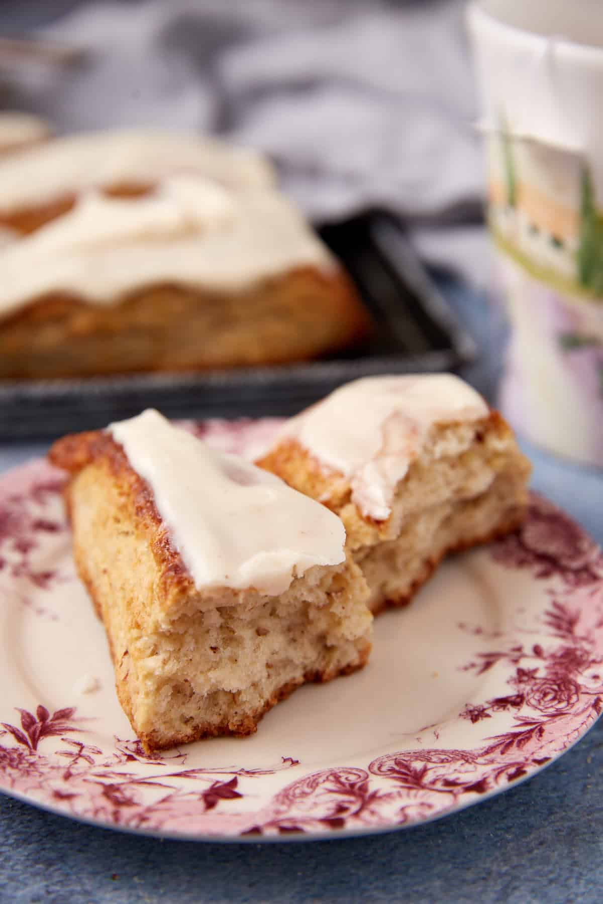 Two pieces of cinnamon roll with white icing sit on a floral-patterned plate, with a tray of more rolls and a cup in the blurred background.