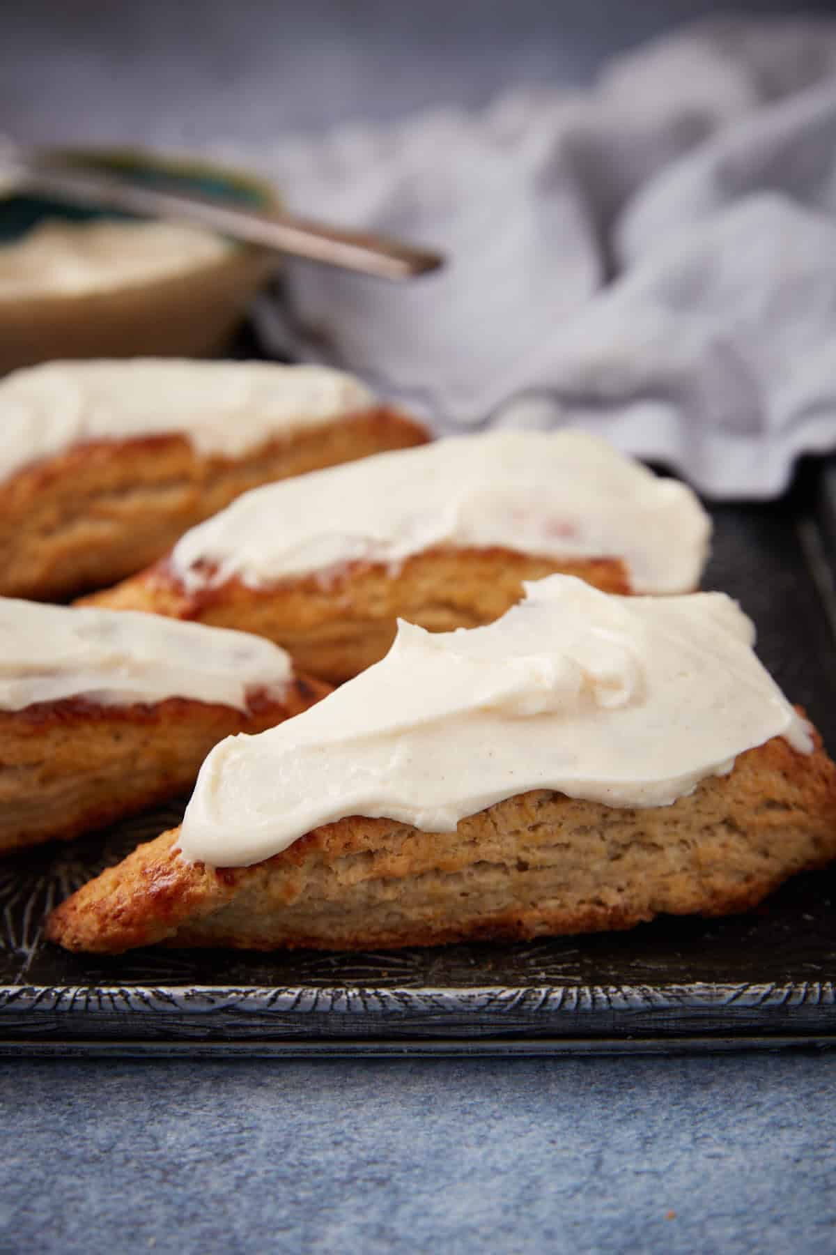 Four triangular scones with creamy white icing are arranged on a dark tray, with a blurred light cloth and a bowl in the background.