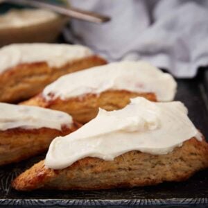 Four triangular scones topped with thick white icing are arranged on a dark tray, with a blurred cloth and a bowl of icing in the background.
