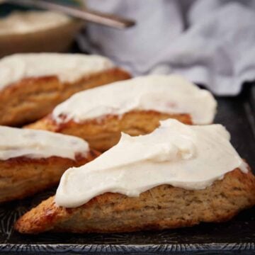 Four triangular scones topped with thick white icing are arranged on a dark tray, with a blurred cloth and a bowl of icing in the background.