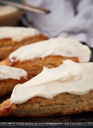 Four triangular scones topped with thick white icing are arranged on a dark tray, with a blurred cloth and a bowl of icing in the background.